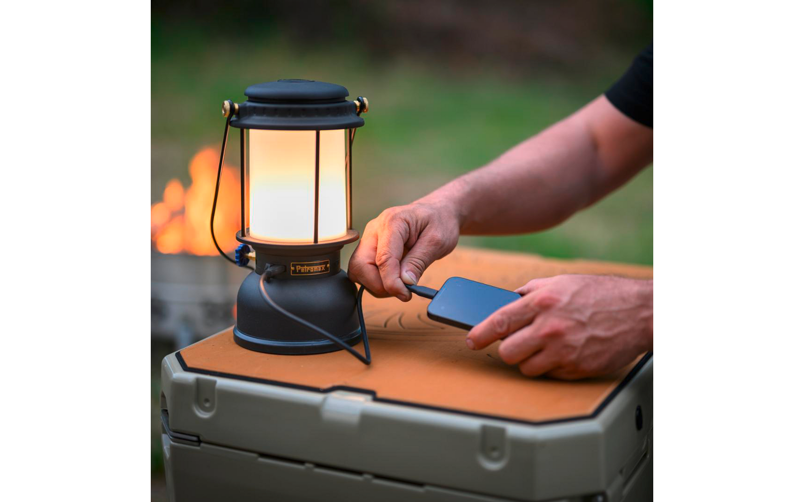 Lanterna da campeggio nera su una cassa di plastica; due mani tengono uno smartphone e avvicinano un cavo alla lanterna; falò sfocato e erba sullo sfondo
