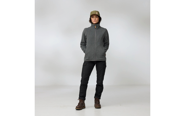 Person steht frontal, Hände in den Taschen, trägt olivgrünen Eimerhut mit kleinem Logo, graue Fleecejacke mit Reißverschluss, schwarze Cargohose und braune Wanderstiefel, heller einfarbiger Hintergrund