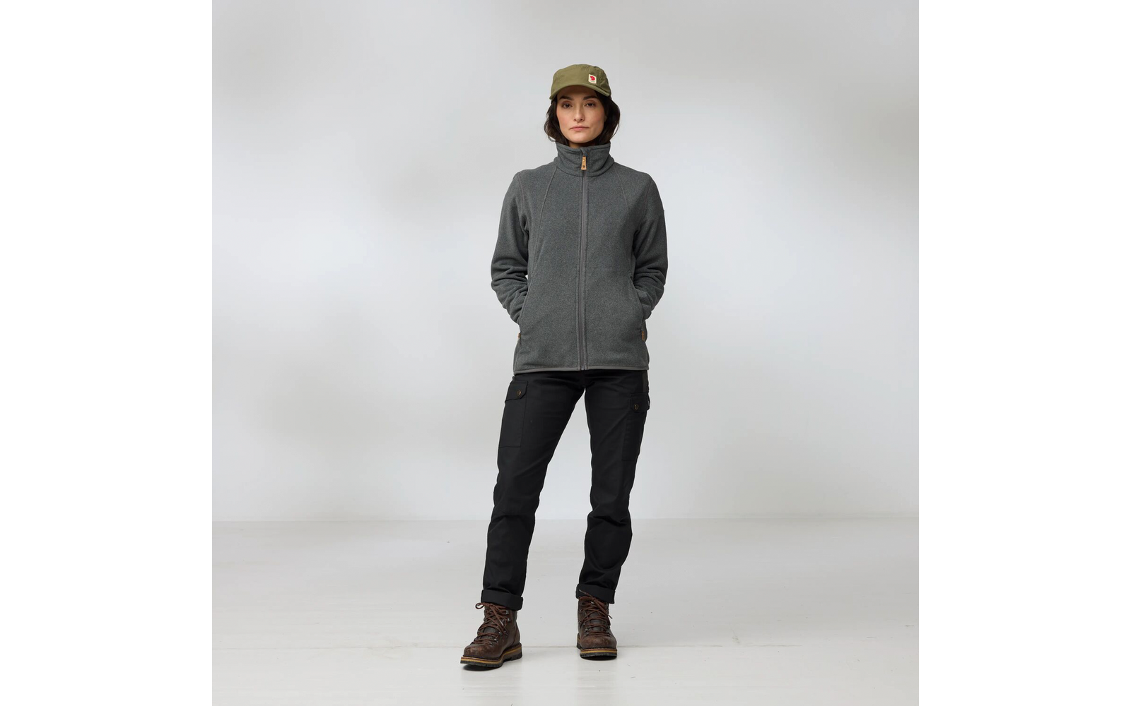 Person steht frontal, Hände in den Taschen, trägt olivgrünen Eimerhut mit kleinem Logo, graue Fleecejacke mit Reißverschluss, schwarze Cargohose und braune Wanderstiefel, heller einfarbiger Hintergrund