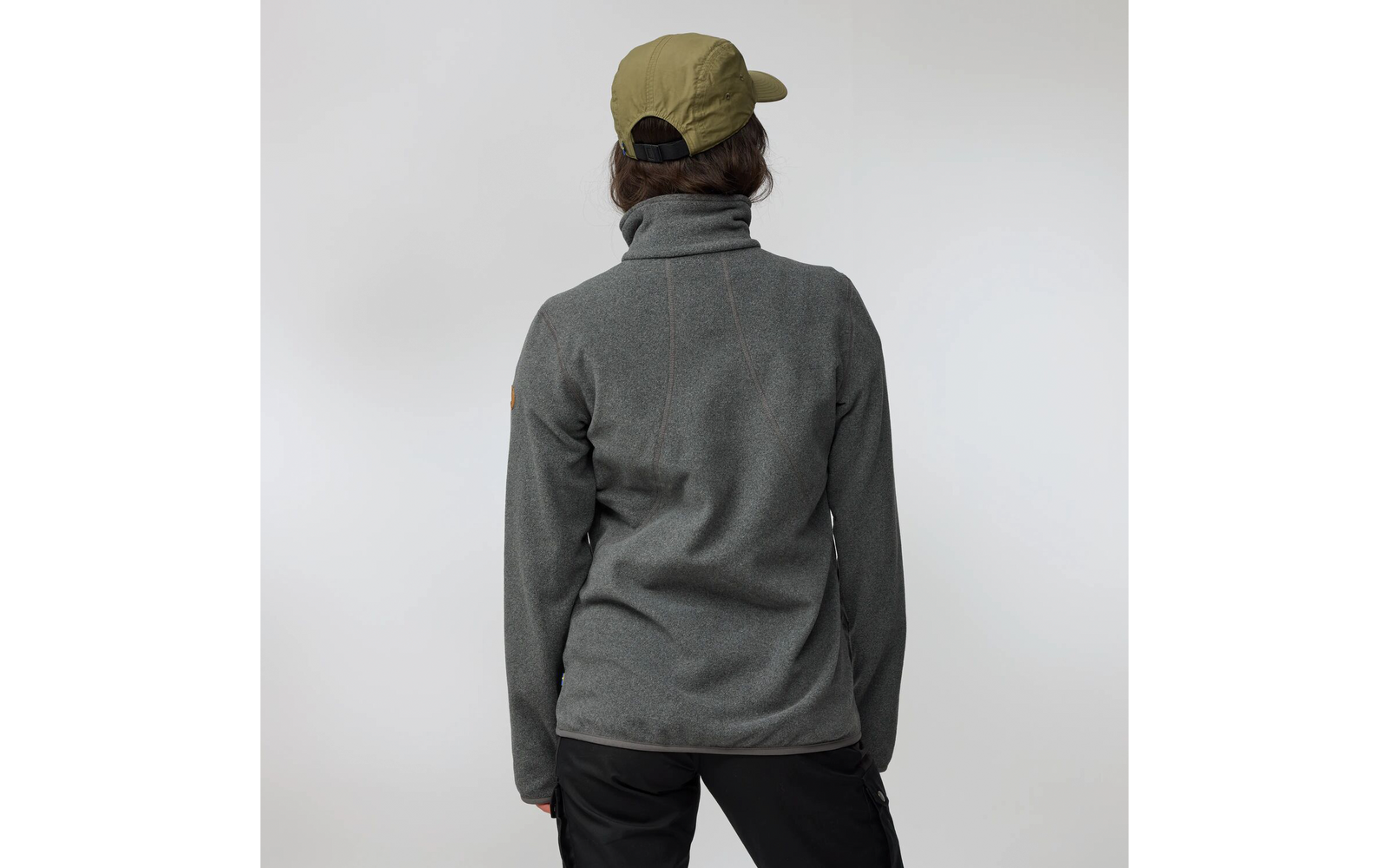 Person von hinten, trägt olivgrüne Baseballkappe, graue Fleecejacke mit hohem Kragen und schwarze Hose vor hellem Hintergrund