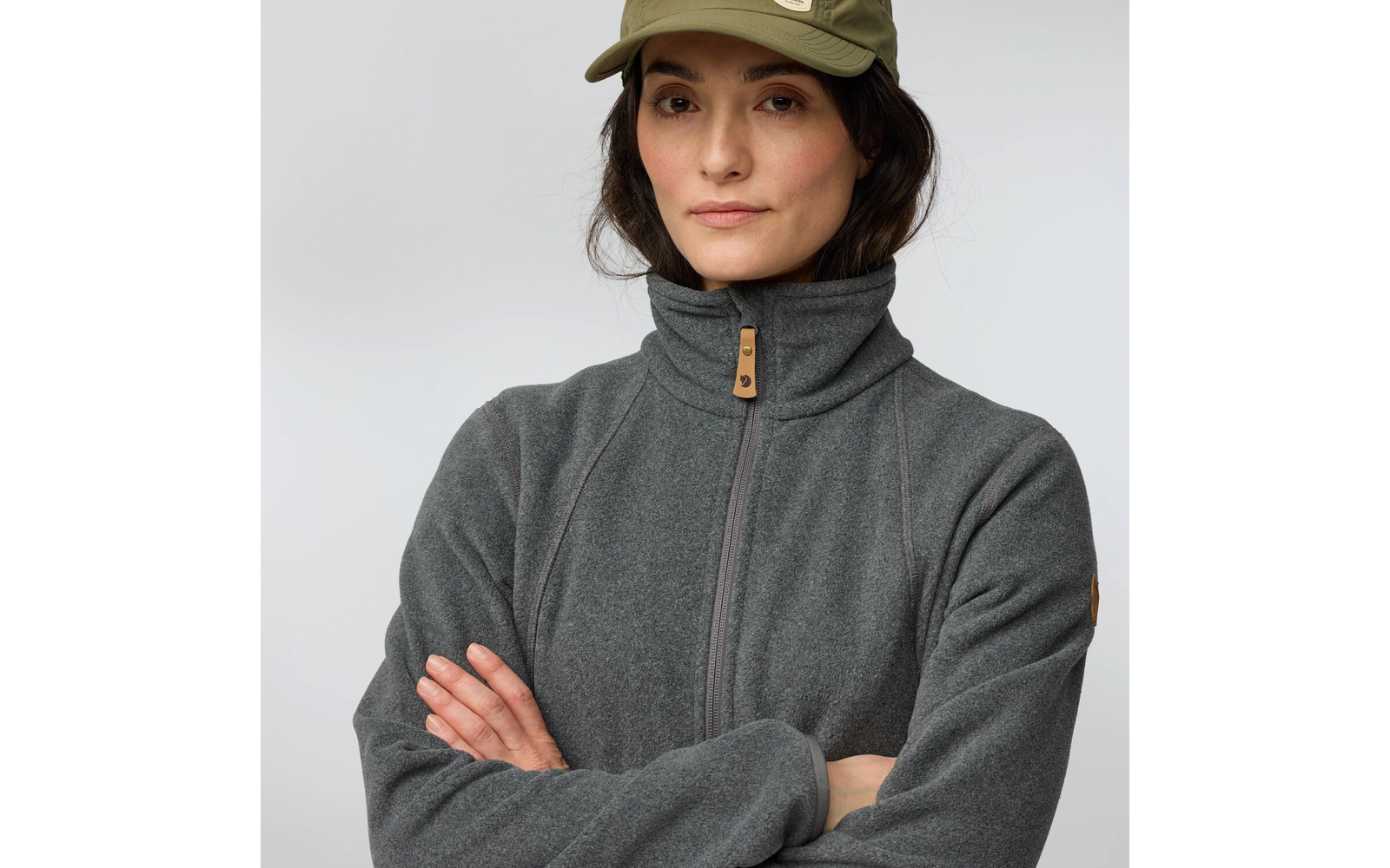 Personne en buste, porte une polaire grise à fermeture éclair et col montant, casquette baseball vert olive, bras croisés, fond uni clair