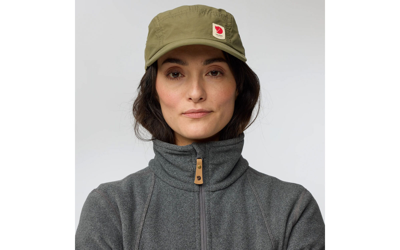 Personne portant une casquette vert olive avec logo, cheveux foncés et veste polaire grise sur fond neutre