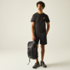 Mann mit dunklen, lockigen Haaren und Schnurrbart, trägt schwarzes T‑Shirt mit kleinem Logo, schwarze Shorts und Sportschuhe, steht und hält einen schwarzen Rucksack an der Seite