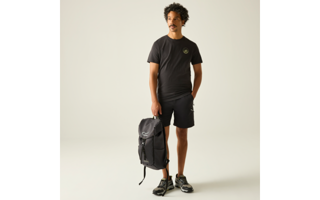 Mann mit dunklen, lockigen Haaren und Schnurrbart, trägt schwarzes T‑Shirt mit kleinem Logo, schwarze Shorts und Sportschuhe, steht und hält einen schwarzen Rucksack an der Seite