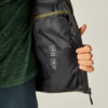 Hand houdt een open jas; zwarte voering met twee witte verzorgingspictogrammen en een klein zakje met olijfgroene bies; groen buitenmateriaal zichtbaar, met een groen shirt en donkere broek eronder