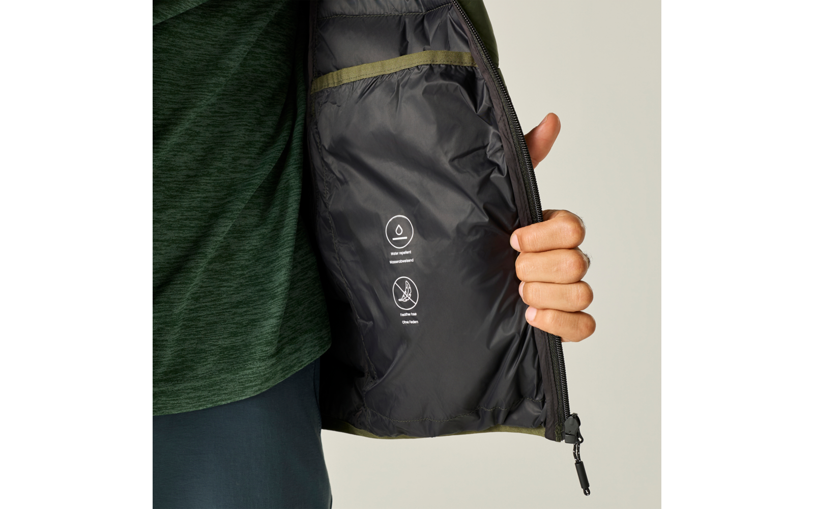 Hand houdt een open jas; zwarte voering met twee witte verzorgingspictogrammen en een klein zakje met olijfgroene bies; groen buitenmateriaal zichtbaar, met een groen shirt en donkere broek eronder