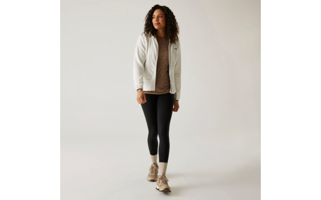 Frau mit langem, lockigem Haar trägt eine helle Jacke, schwarzen Pullover, schwarze Hose und beige Stiefel, steht vor weißem Hintergrund