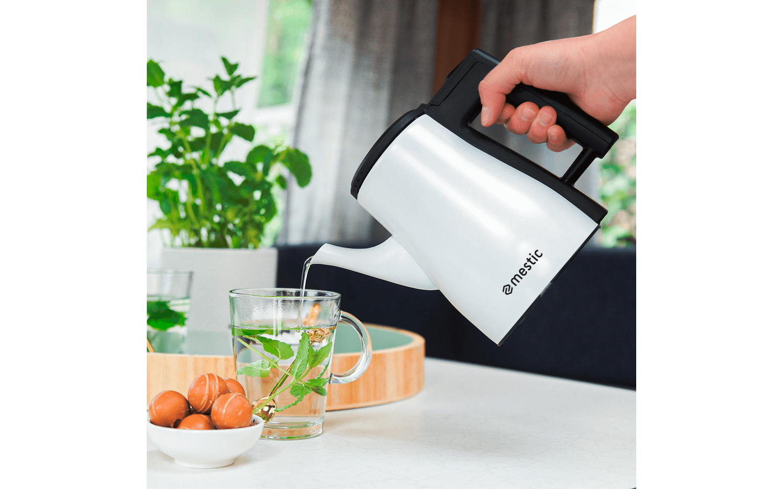 Hand pouring water from a white kettle into a glass cup with a tea bag