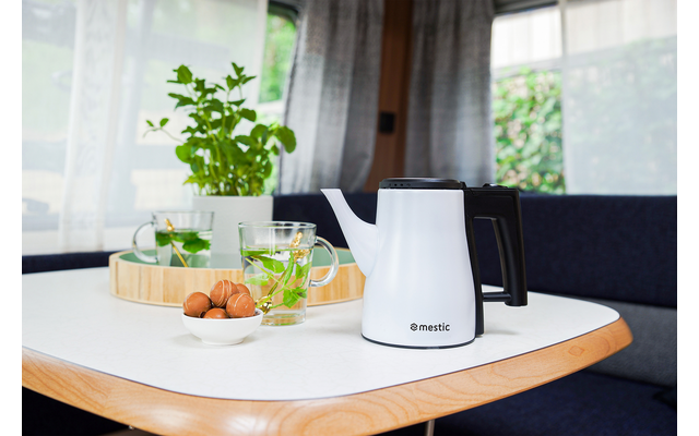 White kettle on a wooden table next to two glasses, one glass with a green drink and a small jug, herbs and windows in the background