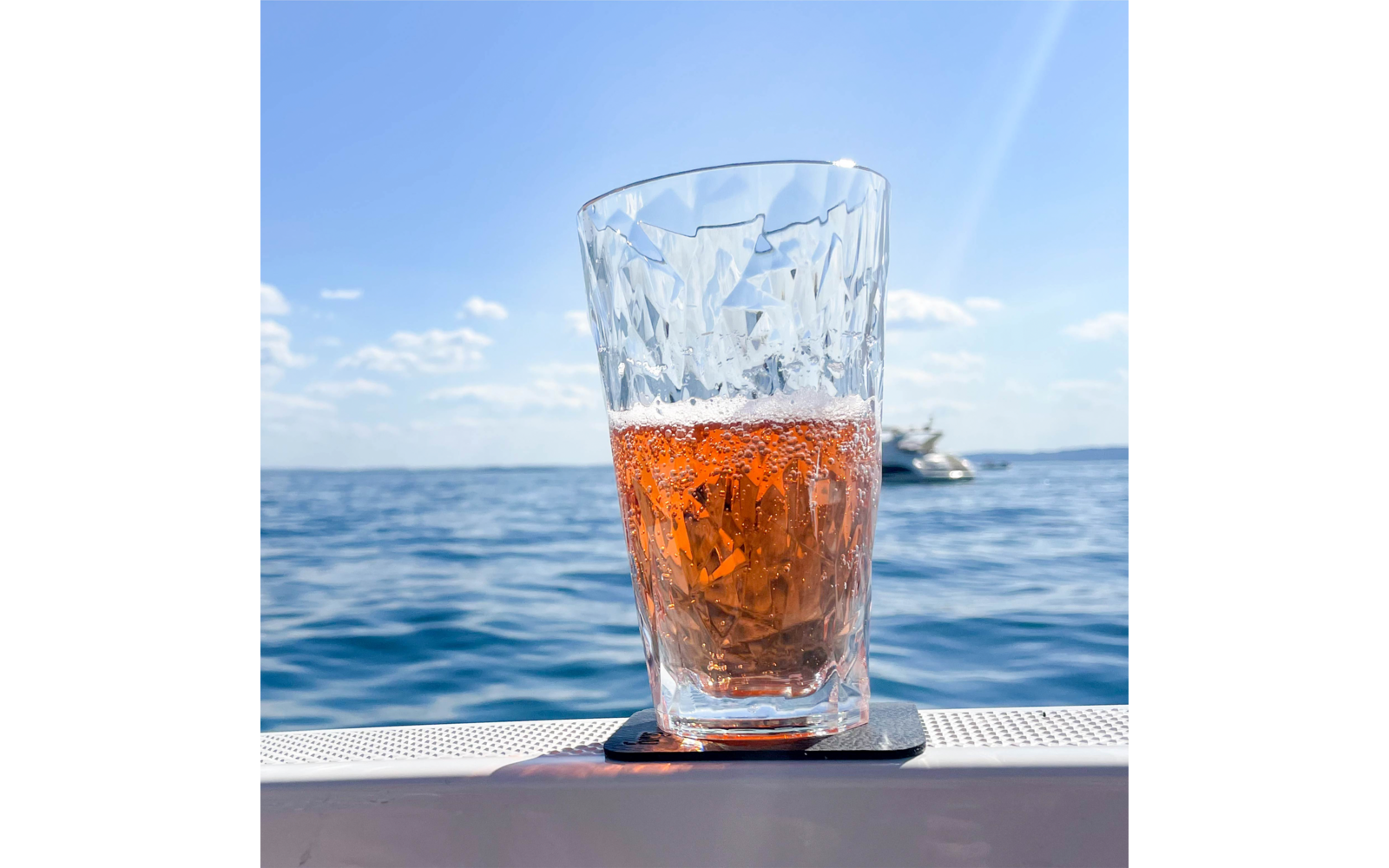 Kristallglas mit rötlich-orangefarbenem Getränk und Bläschen auf einem Untersetzer am Bootsrand; im Hintergrund Meer, ein Motorboot und blauer Himmel