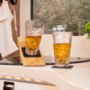 Zwei strukturierte Gläser mit bernsteinfarbener Flüssigkeit auf schwarzen Untersetzern auf einer Arbeitsplatte; links steht ein Glas schräg auf einem Holzbrett. Im Hintergrund ein Fenster mit Bäumen und einer weißen Caravan, links ein weißes Tuch und