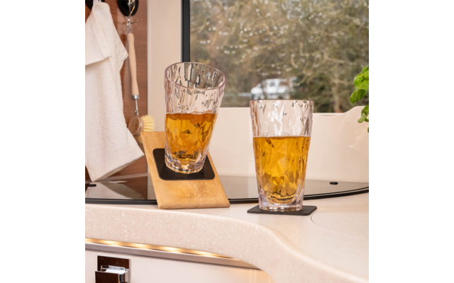 Zwei strukturierte Gläser mit bernsteinfarbener Flüssigkeit auf schwarzen Untersetzern auf einer Arbeitsplatte; links steht ein Glas schräg auf einem Holzbrett. Im Hintergrund ein Fenster mit Bäumen und einer weißen Caravan, links ein weißes Tuch und