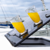Zwei Gläser mit orangefarbener Flüssigkeit auf einer schräg geöffneten Luke eines Segelbootes, im Hintergrund Masten und weitere Boote
