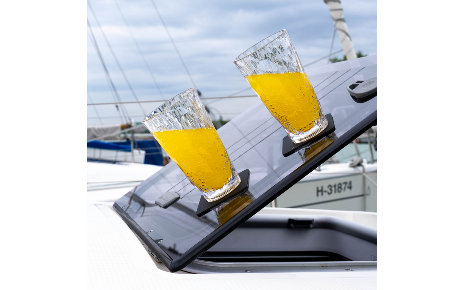 Zwei Gläser mit orangefarbener Flüssigkeit auf einer schräg geöffneten Luke eines Segelbootes, im Hintergrund Masten und weitere Boote