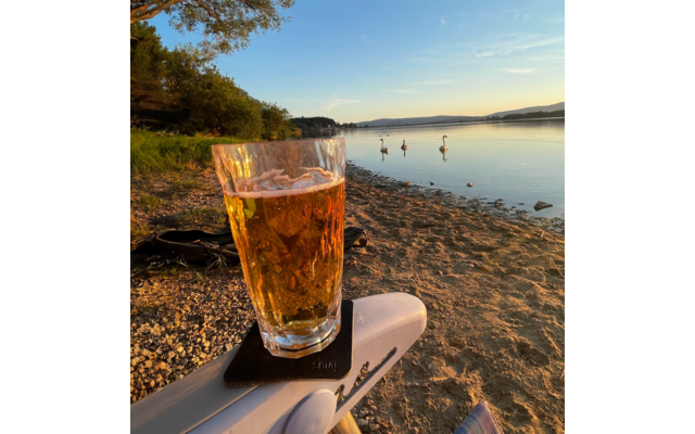 Vordergrund: Glas mit bernsteinfarbener Flüssigkeit und Schaum auf einer weißen Armlehne; Hintergrund: kiesiger Strand, drei auf dem ruhigen See schwimmende Schwäne, Bäume am linken Ufer, Hügel am Horizont, klarer Himmel mit wenigen Wolken