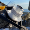 Schwarzes Tablett mit zwei Stielgläsern (orangefarbene und dunkelrote Flüssigkeit) und einer weißen, facettierten Tasse mit Untertasse und schwarzer runder Abdeckung, auf Felsen mit Schneeresten; unscharfer Meereshorizont im Hintergrund