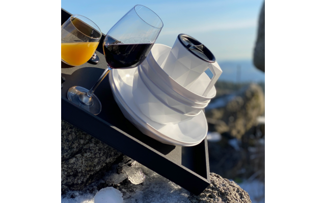 Schwarzes Tablett mit zwei Stielgläsern (orangefarbene und dunkelrote Flüssigkeit) und einer weißen, facettierten Tasse mit Untertasse und schwarzer runder Abdeckung, auf Felsen mit Schneeresten; unscharfer Meereshorizont im Hintergrund