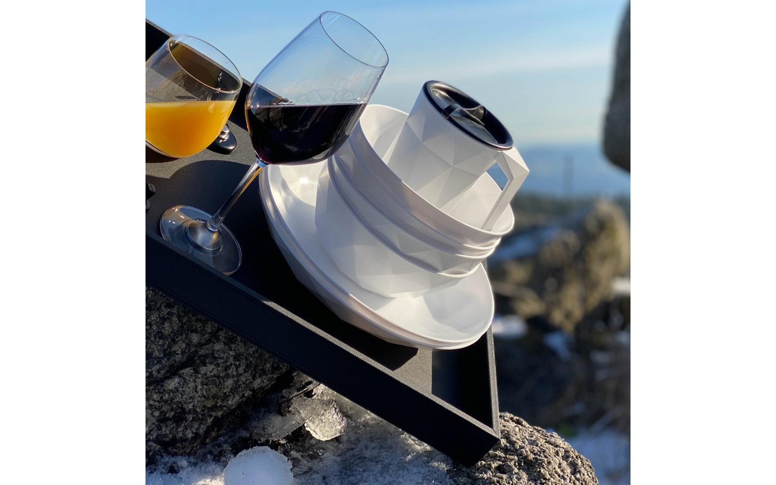 Schwarzes Tablett mit zwei Stielgläsern (orangefarbene und dunkelrote Flüssigkeit) und einer weißen, facettierten Tasse mit Untertasse und schwarzer runder Abdeckung, auf Felsen mit Schneeresten; unscharfer Meereshorizont im Hintergrund