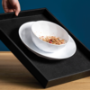 Hand hält schwarzes Tablett mit weißer Schale mit Müsli und weißem Teller, Tablett schräg über Holzfläche vor blauem Hintergrund