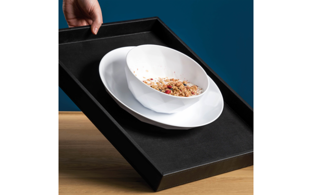 Hand hält schwarzes Tablett mit weißer Schale mit Müsli und weißem Teller, Tablett schräg über Holzfläche vor blauem Hintergrund