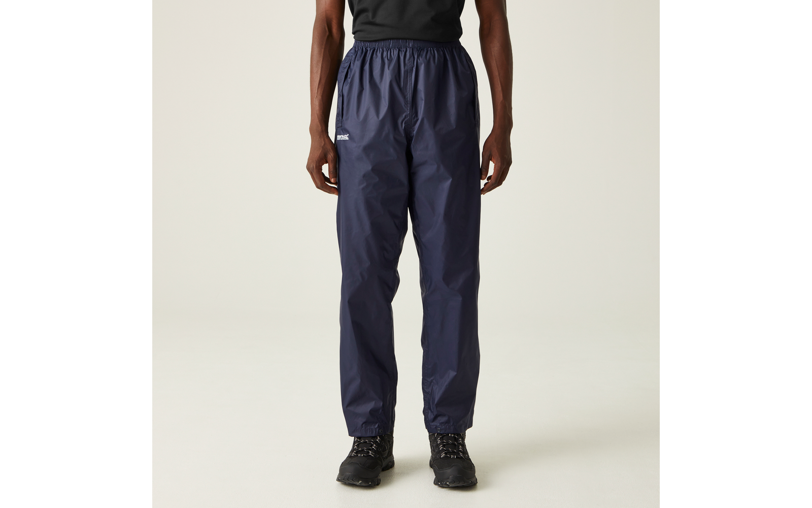 Unterkörper einer stehenden Person von der Taille bis zu den Füßen, dunkelblaue glänzende Regenhose mit elastischem Bund und Bündchen, kleines weißes Logo am rechten Oberschenkel, schwarze Schnürstiefel, einfarbiger hellgrauer Hintergrund