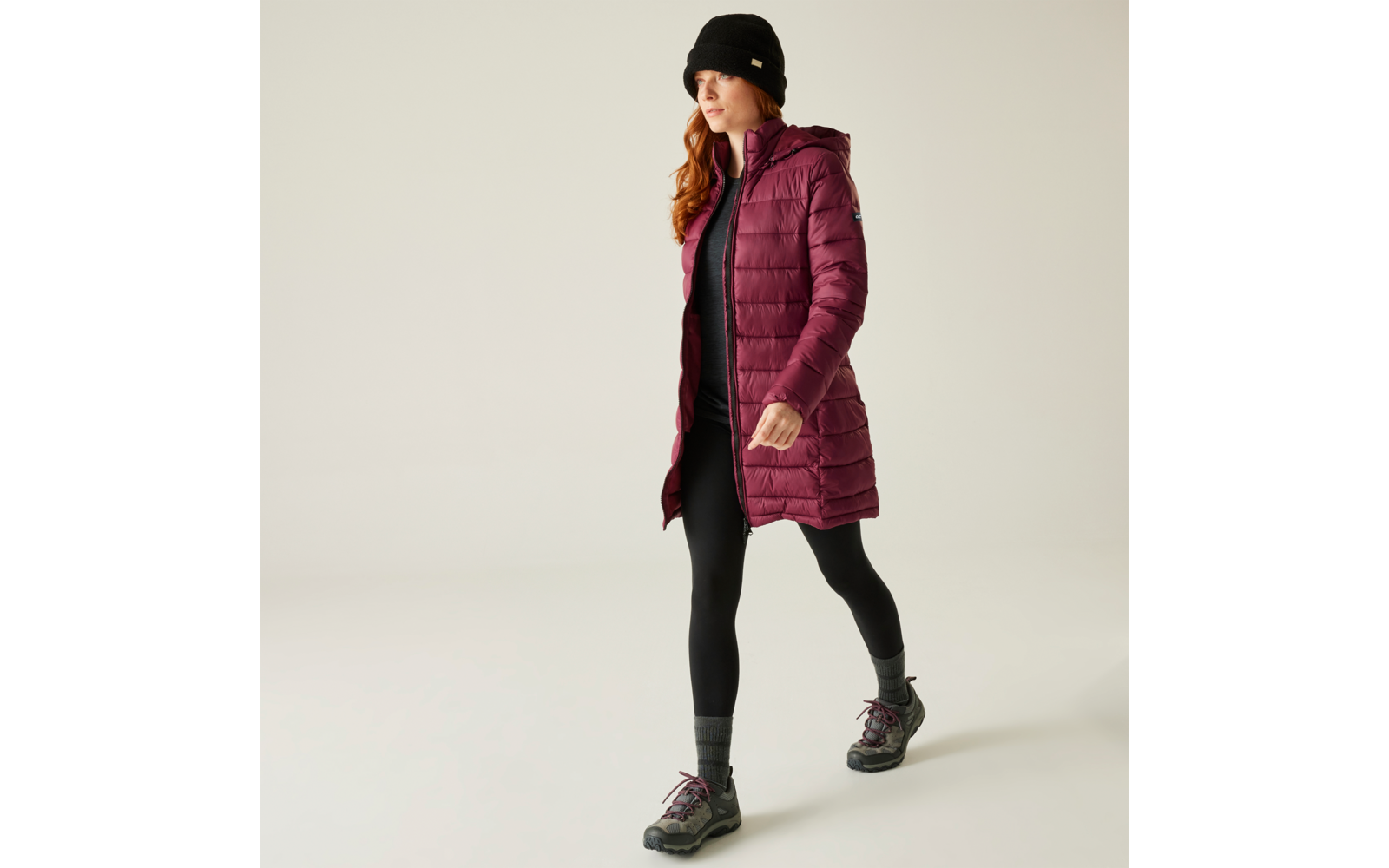 Femme portant un bonnet foncé, un long manteau bordeaux, un pantalon noir et des baskets marchant vers la droite sur fond clair