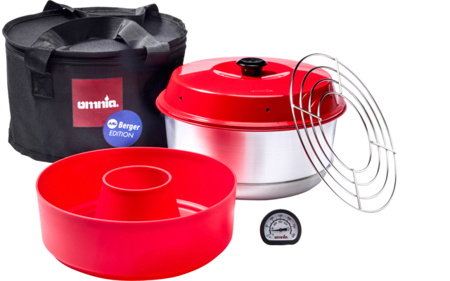 Tapa roja con pomo negro sobre una olla plateada, molde de silicona para bizcocho rojo, rejilla metálica circular, termómetro pequeño y bolsa de transporte negra con los textos 'omnia' y 'Berger Edition'