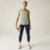 Frau steht frontal, trägt mintgrünes T‑Shirt mit pinkem Aufdruck, dunkelblaue Leggings, beige Sportschuhe und gelbe Socken; hält eine rosa Yogamatte in der rechten Hand vor hellem, neutralem Hintergrund