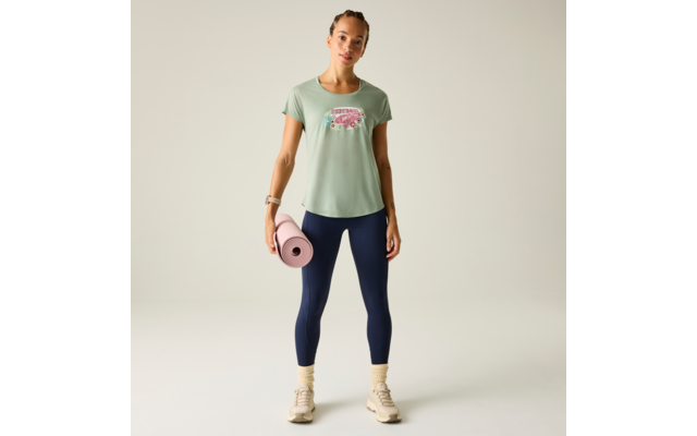 Frau steht frontal, trägt mintgrünes T‑Shirt mit pinkem Aufdruck, dunkelblaue Leggings, beige Sportschuhe und gelbe Socken; hält eine rosa Yogamatte in der rechten Hand vor hellem, neutralem Hintergrund