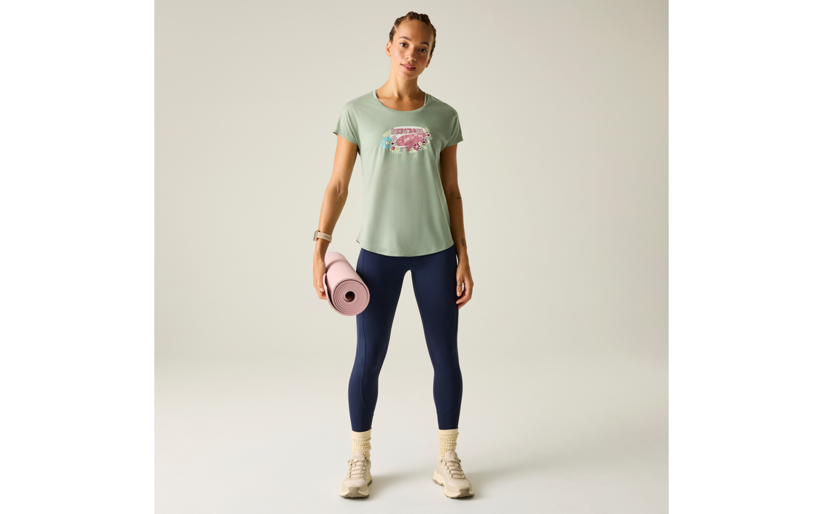 Frau steht frontal, trägt mintgrünes T‑Shirt mit pinkem Aufdruck, dunkelblaue Leggings, beige Sportschuhe und gelbe Socken; hält eine rosa Yogamatte in der rechten Hand vor hellem, neutralem Hintergrund