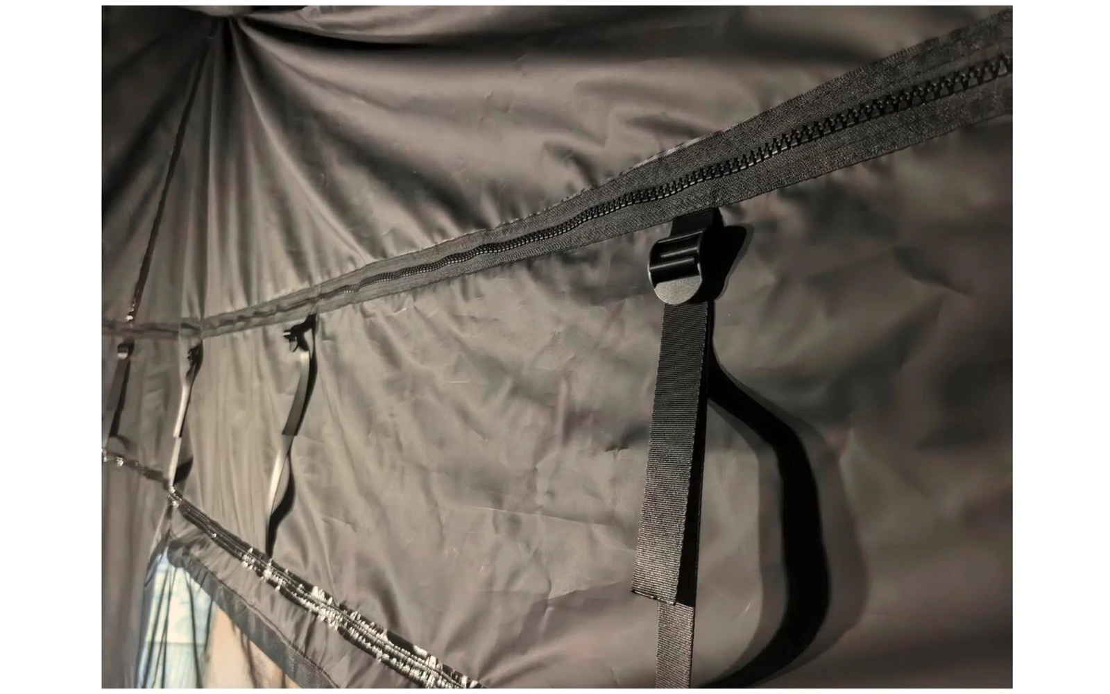 tissu synthétique foncé avec une fermeture éclair horizontale, une sangle noire avec boucle en plastique pendante, coutures et plis visibles, ouverture en tissu clair avec fermeture éclair en bas à gauche