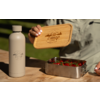 Main aux ongles vernis blanc tient un couvercle en bois avec l'inscription "Camp Life" au‑dessus d'une boîte en acier inoxydable contenant des fraises; une bouteille beige avec le même logo est posée sur une table en bois