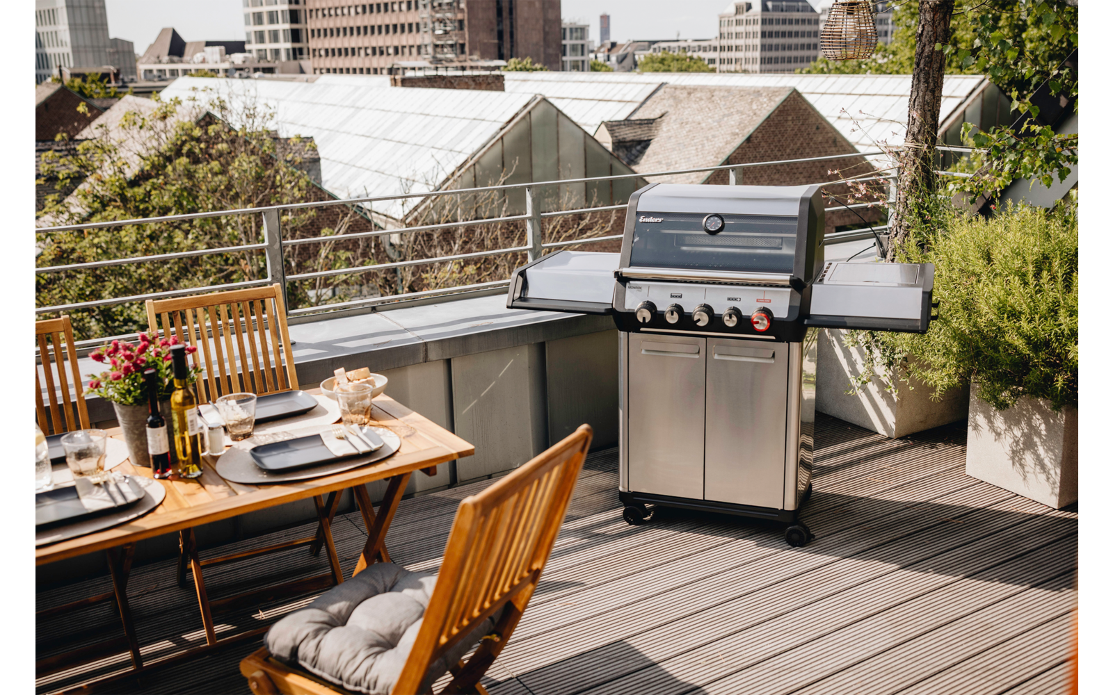 Dachterrasse mit gedecktem Holztisch, Holzstühlen, Edelstahl-Gasgrill, Topfpflanzen und Stadtgebäuden im Hintergrund