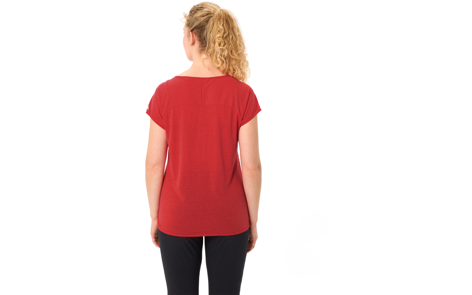 Person von hinten mit blondem, lockigem Pferdeschwanz, rotem T‑Shirt und schwarzer Hose vor weißem Hintergrund