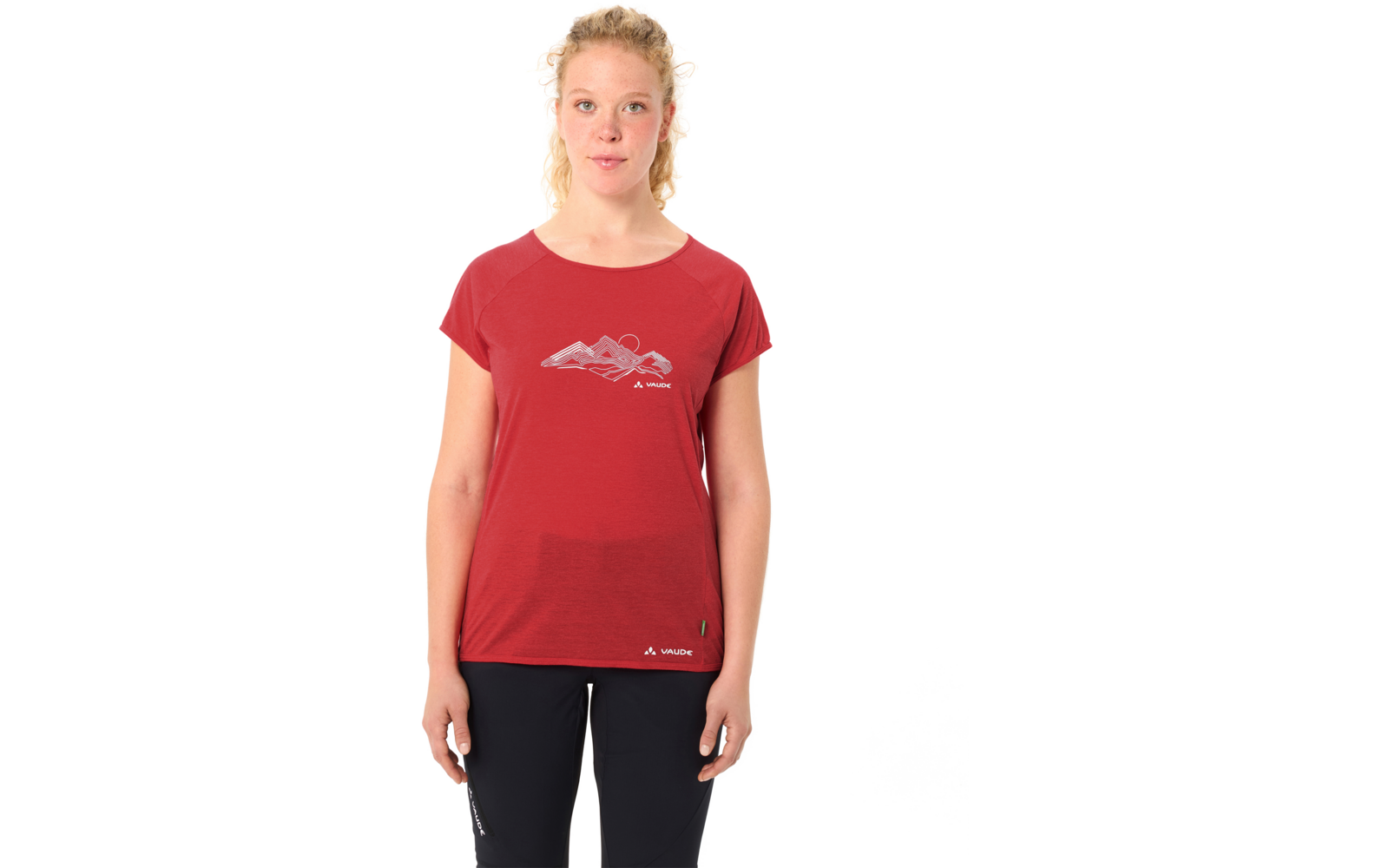 Person steht frontal vor weißem Hintergrund, trägt rotes kurzärmeliges T-Shirt mit Berggrafik und schwarze Hosen, blonde zusammengebundene Haare, Arme seitlich entspannt