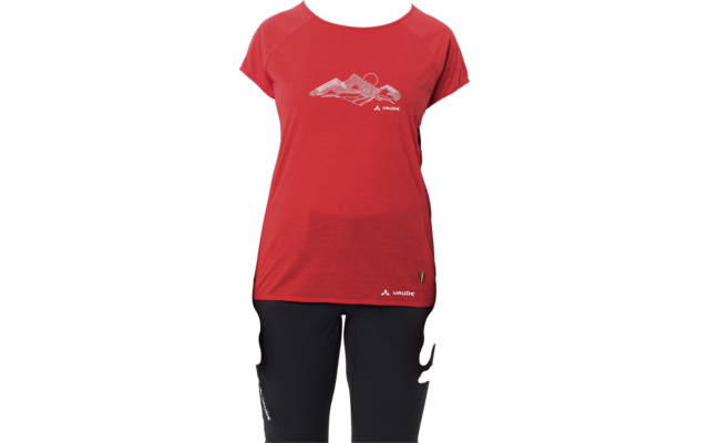 Persona in piedi di fronte alla fotocamera su sfondo bianco, indossa una maglietta rossa a maniche corte con disegno di montagna e pantaloni neri, capelli biondi raccolti, braccia rilassate ai lati