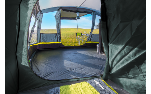 View from inside a tent through the open entrance onto a grassy field; foreground shows tent floor and sleeping bag, outside a camping table, a chair and blue sky