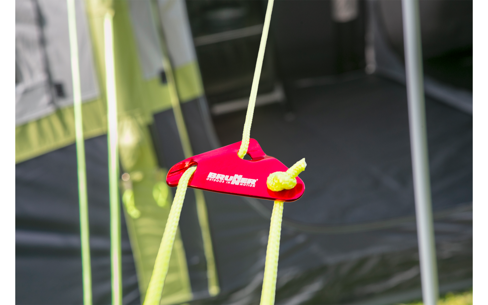 Red plastic rope tensioner piece with two yellow tent cords threaded through; tent fabric and metal poles visible in the background