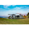 Large gray multi-room tent with front awning and green trim on a grassy field; a camping table with items beside it; mountains, trees with autumn colors and a blue sky with clouds in the background