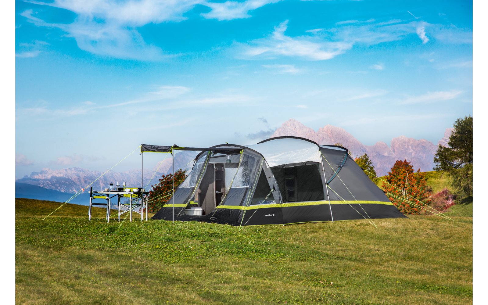 Large gray multi-room tent with front awning and green trim on a grassy field; a camping table with items beside it; mountains, trees with autumn colors and a blue sky with clouds in the background