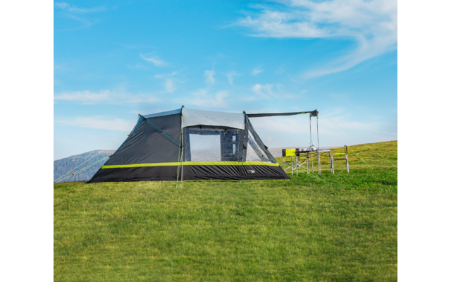 Large gray tent with mesh windows and attached awning on a green hill; camping table and chairs beside it; blue sky with wispy clouds; distant mountains