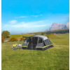 Large gray tent with yellow accents pitched on a green meadow; folding chairs and a table with dishes beside it; trees, rolling hills and mountains in the background under a blue sky