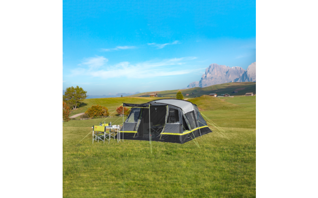 Large gray tent with yellow accents pitched on a green meadow; folding chairs and a table with dishes beside it; trees, rolling hills and mountains in the background under a blue sky
