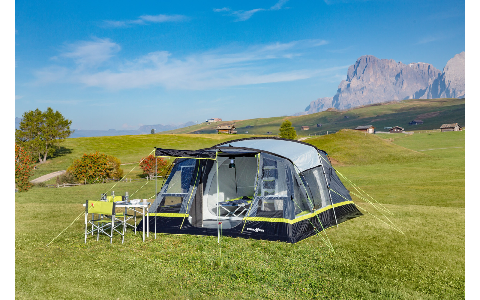 Zelt auf einer grünen Wiese mit Tischen und Stühlen, im Hintergrund Berge und blauer Himmel