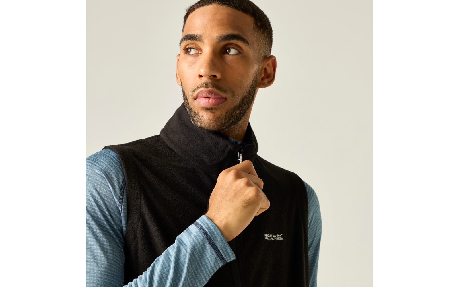Persoon trekt aan de rits van een zwart fleecevest, draagt een blauw shirt met lange mouwen, kijkt naar links, lichte achtergrond