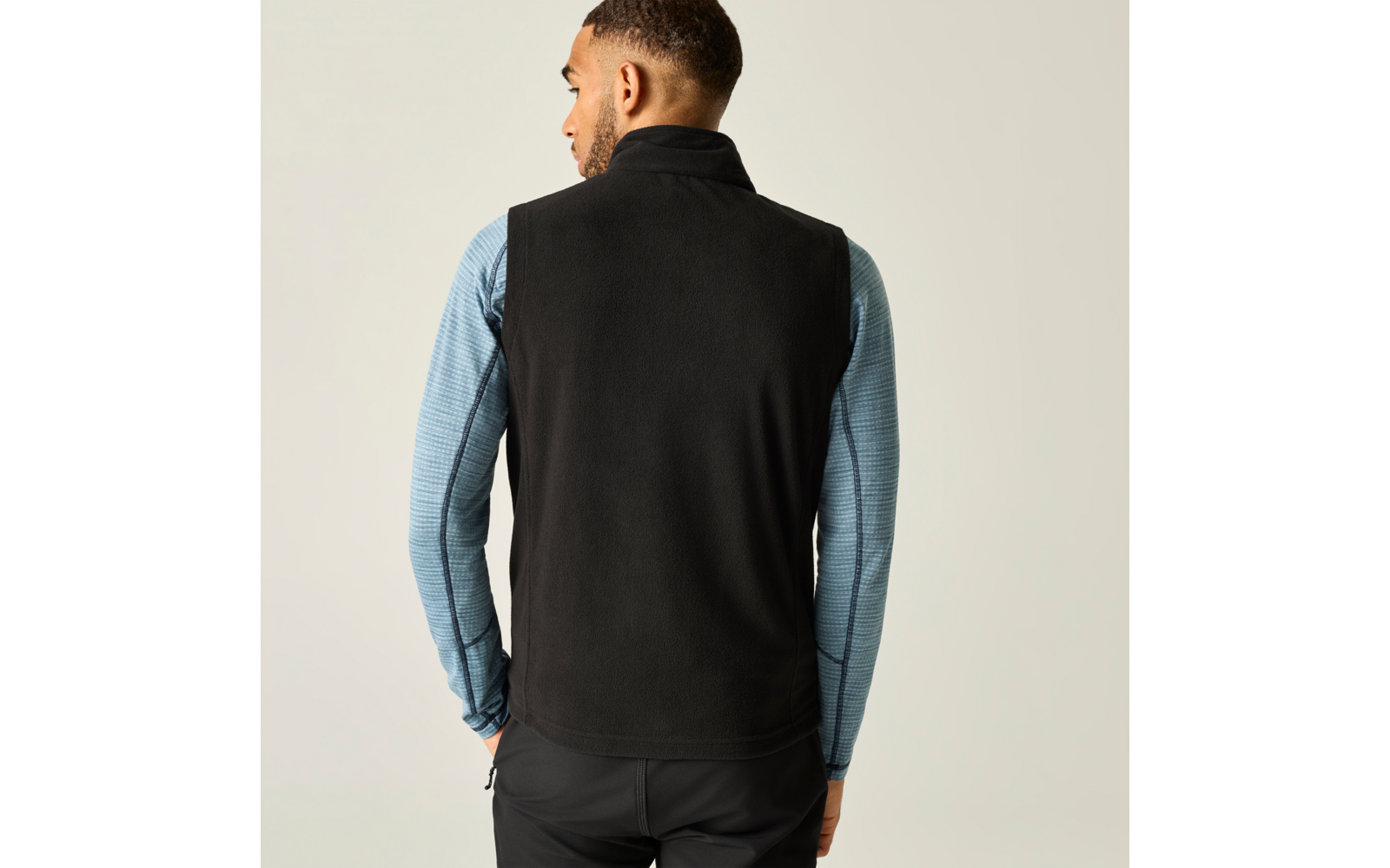 Persoon van achteren, draagt zwart mouwloos fleecegilet over lichtblauw shirt met lange mouwen, voor een lichte achtergrond