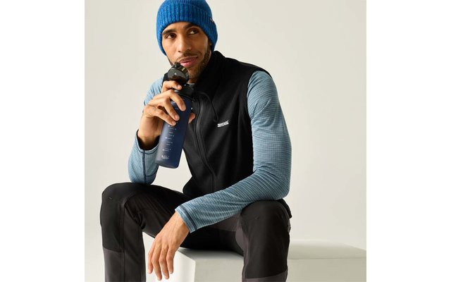 Man zit op een wit blok, houdt een donkerblauwe drinkfles dicht bij zijn mond, draagt een blauwe gebreide muts, zwart vest over een blauw gestreept shirt met lange mouwen en zwarte broek