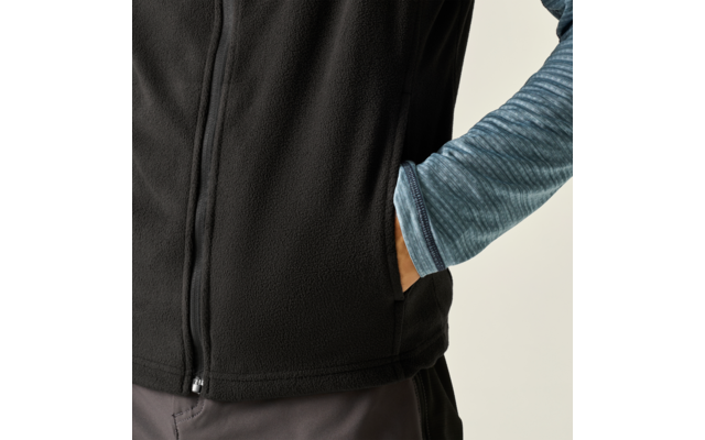 Bovenlichaam van een persoon met een zwart fleecevest over een blauw gemêleerd shirt met lange mouwen, rechterhand in de zak van het vest