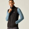 Persoon tegen lichte achtergrond draagt zwarte mouwloze fleece-vest met rits over een blauw, langmouwig shirt; rechterhand bij de rits, linkerhand in de zak, kijkt naar rechts