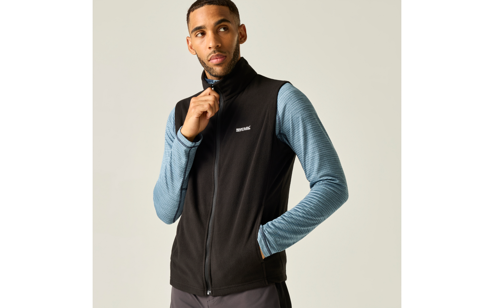 Persoon tegen lichte achtergrond draagt zwarte mouwloze fleece-vest met rits over een blauw, langmouwig shirt; rechterhand bij de rits, linkerhand in de zak, kijkt naar rechts
